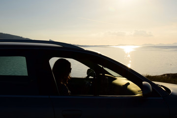A young woman sits in a car by the sea and watches the sunset.