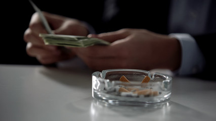 Cigarette ashtray, person counting money on background, illegal business deal