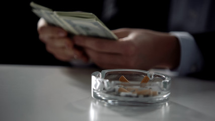 Obraz premium Cigarette butts in ashtray, man counting money on background, tobacco industry