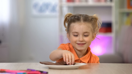 Naklejka premium Female kid secretly taking chocolate from white plate on table, sugar overdose