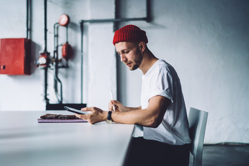 Side view of Caucasian male in trendy outfit sitting at office desktop and using modern cellphone gadget for checking information while working with project, millennial man typing text message