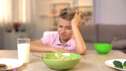 Obraz na płótnie Canvas Unhappy boy looking oatmeal with disgust, unappetizing food, healthy breakfast