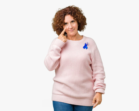 Middle ager senior woman wearing changeable blue color ribbon awareness over isolated background Pointing to the eye watching you gesture, suspicious expression