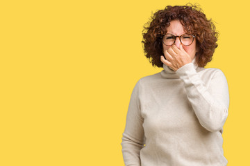 Beautiful middle ager senior woman wearing turtleneck sweater and glasses over isolated background smelling something stinky and disgusting, intolerable smell, holding breath with fingers on nose. 