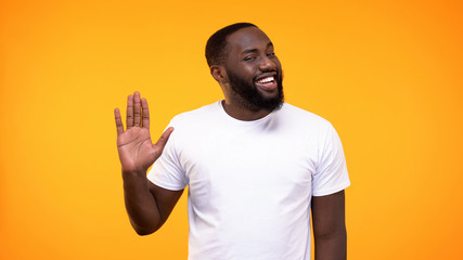 Affable young black man showing palm, waving hand neighborly yellow background