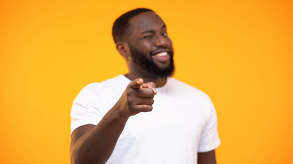 Young black man pointing finger on camera and smiling against yellow background
