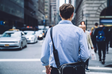 Back view of intelligent male economist with trendy leather briefcase walking to business meeting in financial district, clever professional proud ceo in blue t shirt going to destination in downtown