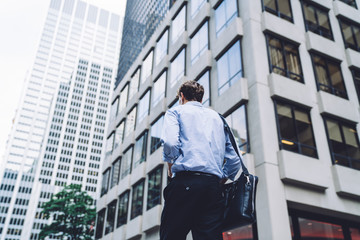 Back view of trendy businessman with leather briefcase walking at city street in downtown, stylish male entrepreneur formally dressed going to meeting in financial district of megalopolis