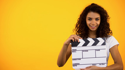 Smiling young female holding clapper board on orange background, cinematography