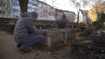 Young man sitting near grave on ancient cemetery praying, mourning for relatives