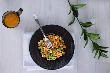 Frozen fried vegetables on black plate. Vegan breakfast with juice. Vegetables food mix on white wooden background with plant.