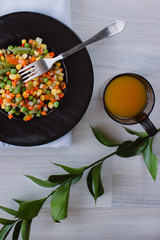 Frozen fried vegetables on black plate. Vegan breakfast with juice. Vegetables food mix on white wooden background with plant.