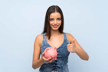 Young woman over isolated blue wall holding a big piggybank