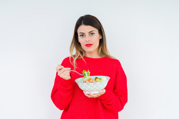 Young woman eating fresh healthy salad over isolated background with a confident expression on smart face thinking serious