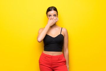 Young woman over isolated yellow background covering mouth with hands