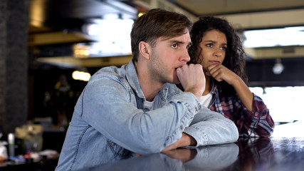 Multiethnic couple watching sport match on big screen in bar, upset with results