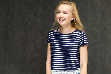 Portrait to the waist of a pretty interesting fashionable blonde girl on a gray background in a striped blue t-shirt. Standing in front of the camera, smiling, showing hands. Shows a lot of emotions.