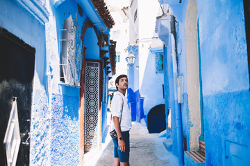 Pensive indian hipster guy standing at antique blue street in old city of Morocco enjoying summer trip for exploring new country, handsome man with retro camera looking around during sightseeing