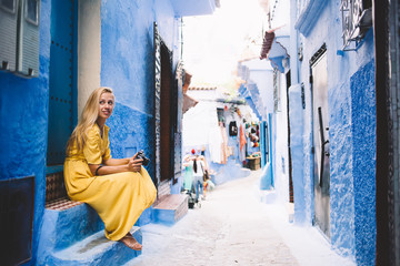 Attractive hipster girl sitting near blue wall in colorful street - Morocco thinking about unesco heritage, young female in yellow dress with camera taking rest in berber medina on vacation