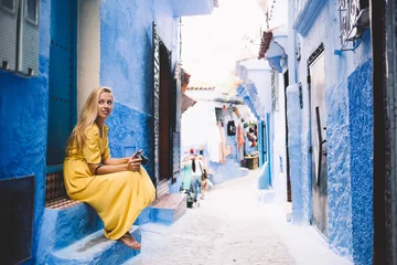 Papier Peint photo Maroc Attractive hipster girl sitting near blue wall in colorful street - Morocco thinking about unesco heritage, young female in yellow dress with camera taking rest in berber medina on vacation