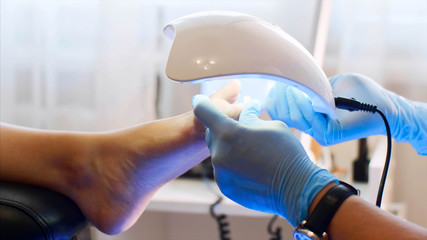 Professional pedicure is beauty salon. Pedicurist woman in gloves is holding UV lamp above the foot. She is drying shellac on client toes, foot closeup.