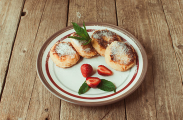 Fototapeta na wymiar Gourmet breakfast - cottage cheese pancakes, cheesecakes, cottage cheese pancakes with strawberries, mint and powdered sugar in a white plate. Useful dessert on a wooden table in rustic style. Selecti