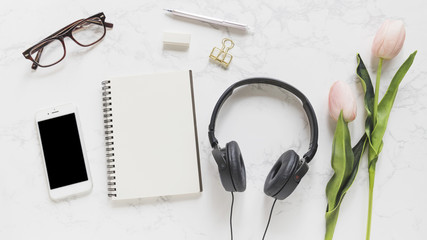Mobile phone; eyeglasses; notebook; stationery; headphone and pink tulips on marble backdrop