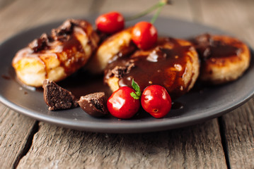 Gourmet breakfast - cottage cheese pancakes, cheese cakes, cottage cheese pancakes with cherries and chocolate in a brown plate. Useful dessert on a wooden table in rustic style. Selective focus.