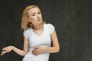 Waist up portrait of pretty interesting fashionable blonde girl on gray background in white t-shirt. Standing in front of the camera, smiling, showing hands. Shows a lot of emotions.