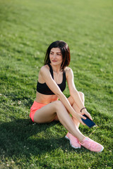 Fototapeta premium Young sporty joyful woman brunette in shorts and top sitting on the grass football field stadium and listens to music in earphones, she gets pleasure, relaxes. Sport and healthy lifestyle