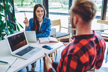 Positive male and female hipsters planning future event with friends from university using textbook and organiser application on laptops, happy freelance colleagues collaborating on netbooks