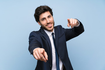 Businessman over isolated blue wall points finger at you while smiling