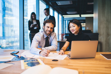 Portrait of cheerful successful male and female colleagues smiling at camera during cooperation process in office room, happy man and woman in formal wear sitting at desktop with modern laptop