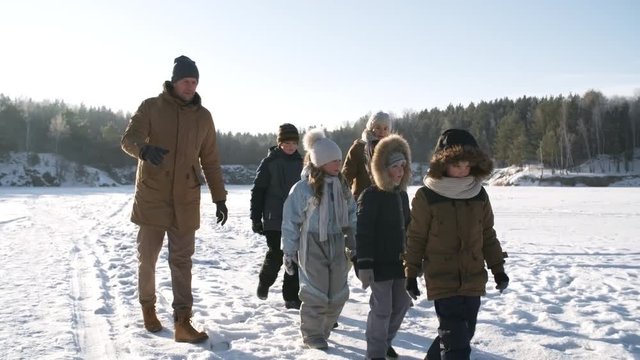 Following shot of active large young family with five children strolling in snow over frozen surface of lake in bright daylight, father talking to kids and mother smiling happily
