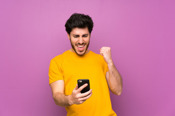 Handsome over isolated purple wall with phone in victory position