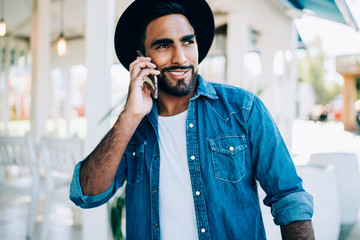 Happy hipster guy dressed in stylish apparel enjoying positive smartphone conversation via app, cheerful Spanish man making international phone call during leisure time connected to roaming