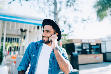 Spanish hipster guy in stylish apparel enjoying positive cell conversation during leisure on urban...