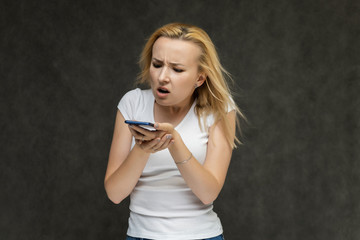 Fototapeta premium Waist up portrait of pretty interesting fashionable blonde girl on gray background in white t-shirt with smartphone. Standing in front of the camera, smiling, showing hands. many emotions.