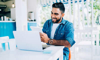 Successful man reading information from textbook during time for e learning excited with interesting idea for university essay, positive Turkish man sitting at cafeteria terrace and preparing to exams