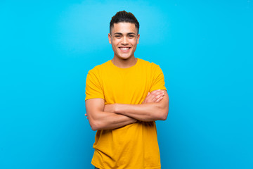 Young man with yellow shirt over isolated blue background keeping the arms crossed in frontal...