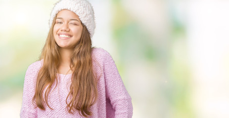 Young beautiful brunette woman wearing sweater and winter hat over isolated background winking...
