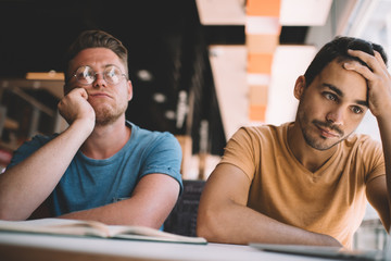 Bored male student in spectacles for vision correction pondering on information for startup project while exhausted colleague with headache sitting near, disappointed men unhappy with overtime