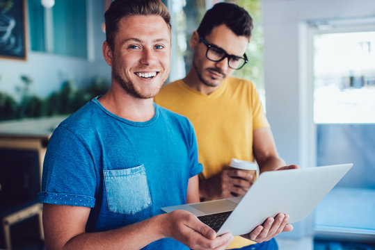 Half length portrait of cheerful Caucasian male it professional holding laptop computer in hand and smiling at camera while colleague standing near and checking text information on website