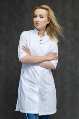 Portrait of a knee-high pretty interesting beautiful girl blonde doctor on a gray background in a white medical coat. Standing in front of the camera, smiling, a lot of emotions.