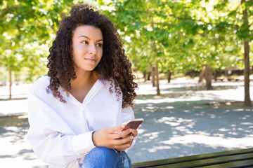 Content pretty young woman using smartphone on bench in park. Beautiful lady wearing blouse and sitting with green trees in background. Communication and nature concept. Front view.