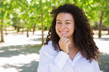 Happy pretty curly-haired lady posing at camera in park. Beautiful young lady wearing blouse and looking at camera with green trees in background. Woman portrait concept. Front view.