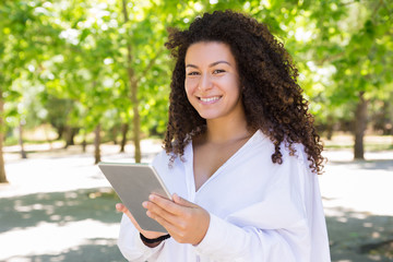 Fototapeta premium Happy pretty young woman browsing on tablet in park. Beautiful lady wearing blouse and looking at camera with green trees in background. Technology and nature concept. Front view.