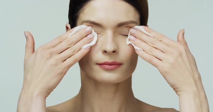 Close up of the Caucasian young pretty woman removing makeup with cotton pads rom eyes and looking to the camera on the white background.
