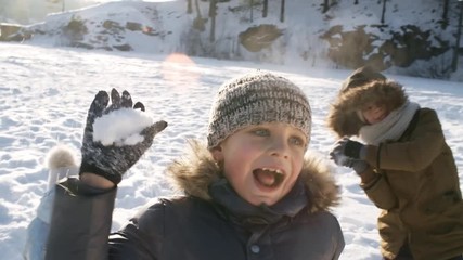 Chest-up shot with lens flare of young boy, wearing hooded jacket and stripy woolly hat, enjoying snowball play fight with siblings and parents during family countryside walk on sunny cold winter day - Powered by Adobe