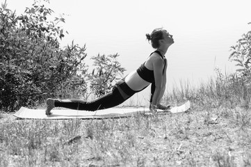 Cute young girl performs yoga asanas on the bank of a river, lake or sea on a sunny summer morning. Diet, exercise and yoga in nature, bw